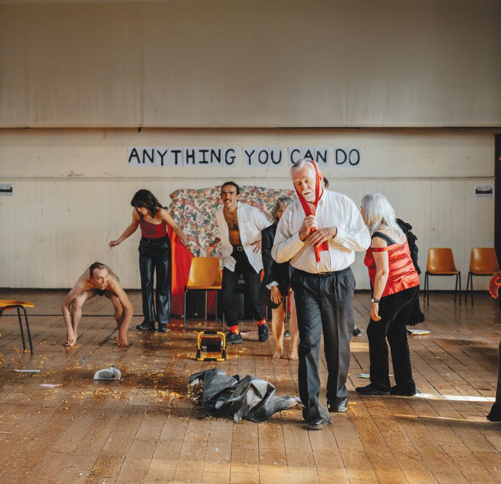 Publicity image for Pony Cam's 'Anything You Can Do' which opens HotHouse Theatre's 2025 season. A fair-skinned older man in suit pants and a white collared shirt stands with a red tie wrapped over his head like a scarf. Behind him is a chaotic scene: the considerably younger members of Pony Cam stand, crouch and run across a wooden floor littered with what could be food scraps; two older, grey and white haired women are also visible in the background.