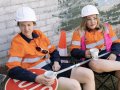 Two female road workers dressed in white helmets and orange hi-vis jackets sit in folding chairs. They nurse cups of tea and a Stop sign is drapped across their laps. A traffic cone is visible behind them.