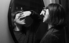 A black and white photo of a woman facing a round mirror with one hand covering her face.